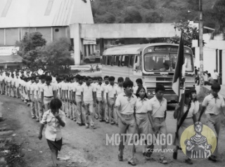 Alumnos de la Secundaria Técnica durante un desfile, marchando frente a la ermita del barrio El Nido, se observan los caminos de terracería, los autobuses de la época y la entrada del ingenio Central Motzorongo.
