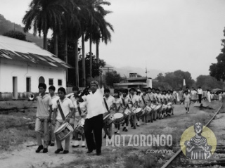 Alumnos de la Secundaria Técnica durante un desfile, al frente de la banda de guerra el profesor Jesús Godínez Olmedo, al fondo la tienda grande, la gerencia y la nueva estación del ferrocarril.